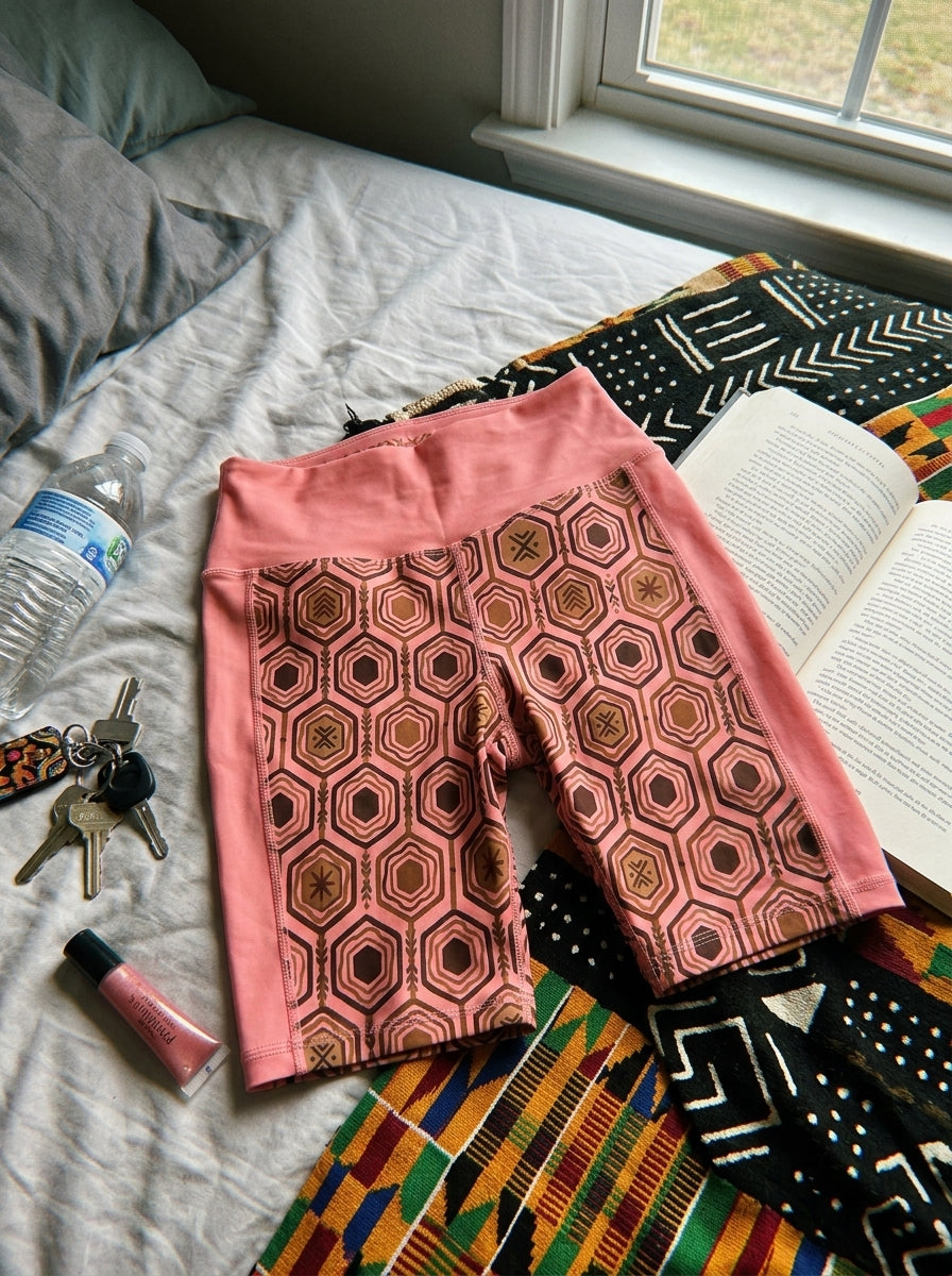 AnanseNova™ Biker Shorts in Adaeze Pink, flat lay on bed with kente cloth and accessories — SunBorn