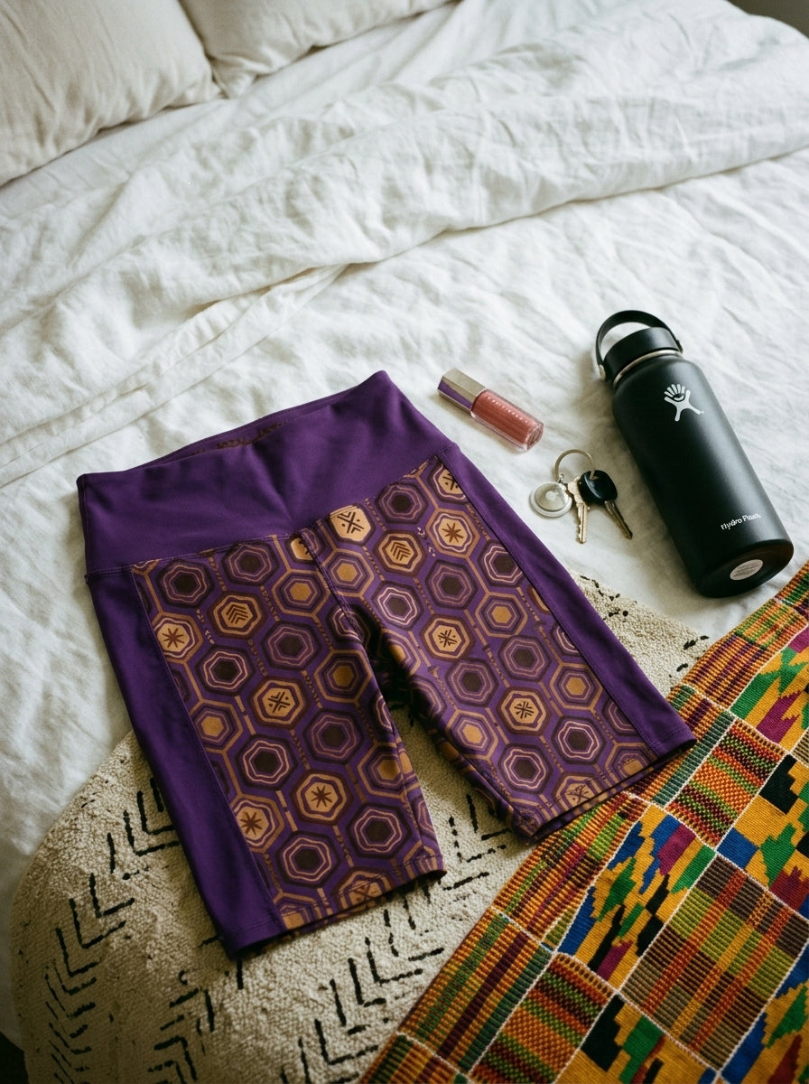 AnanseNova™ Biker Shorts in Ugo Purple, flat lay on bed with kente cloth and water bottle — SunBorn