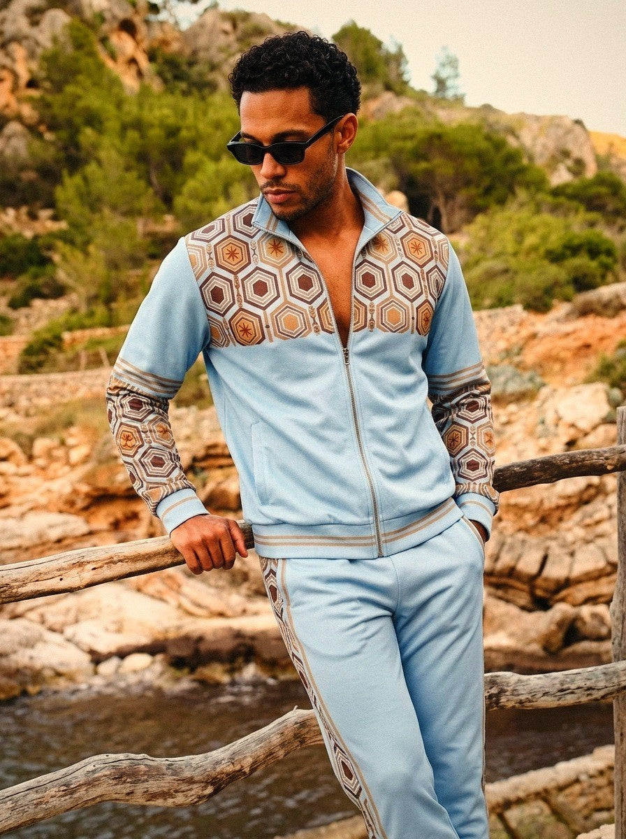 AnanseNova™ Track Jacket + Joggers Set in Igwe Blue, lifestyle shot: model in sunglasses leaning on a wooden rail above a rocky Mediterranean cove