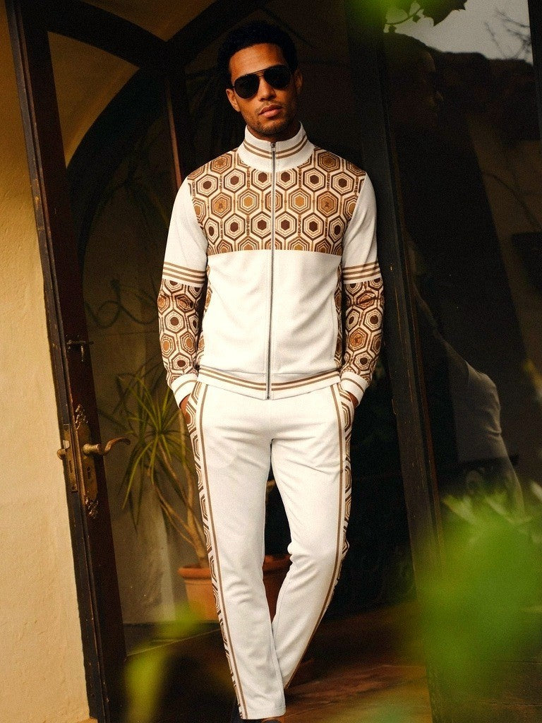 AnanseNova™ Track Jacket + Joggers Set in Ocha White, lifestyle shot: model in sunglasses leaning in a doorway, white track jacket with brown and gold hexagon print yoke and sleeves, matching joggers with hex side stripes