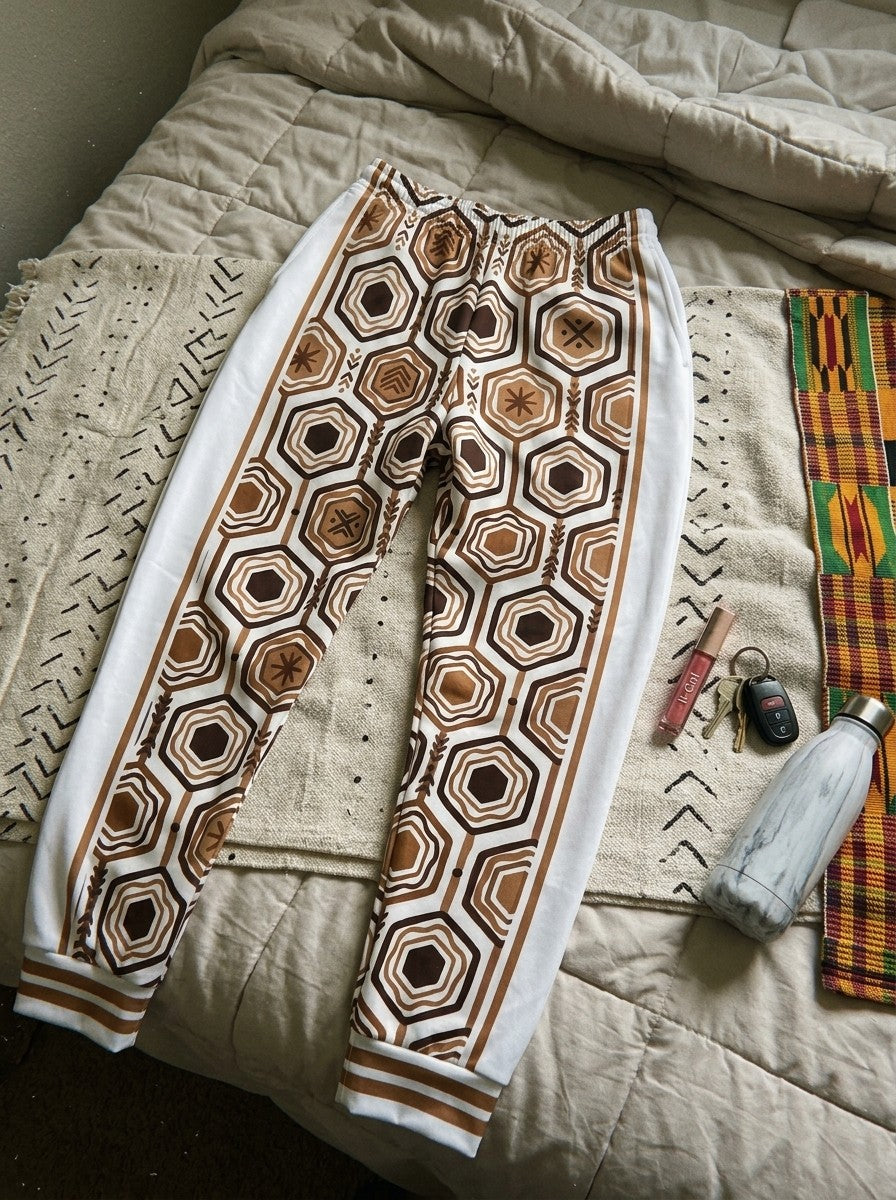 AnanseNova™ Women's Joggers in Ocha White laid on a bed with mudcloth and kente throws, keys, lip gloss and a water bottle