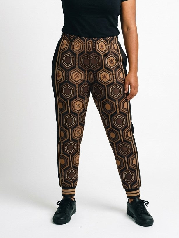 AnanseNova™ Women's Joggers in Oji Black, front view on model, full-front bronze and brown hexagon print with black side panels, bronze side stripes and striped cuffs | SunBorn