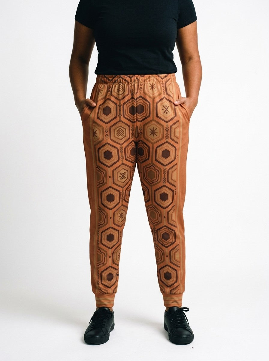 AnanseNova™ Women's Joggers in Sango Orange, front view on model, full-front brown and bronze hexagon print with orange side panels, bronze side stripes and striped cuffs | SunBorn