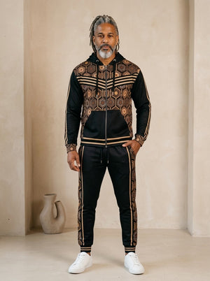AnanseNova™ Zip Hoodie + Joggers Set in Oji Black, front view on model: black zip hoodie with brown and gold hexagon print panels and gold sleeve stripes, matching black joggers with hex side stripes