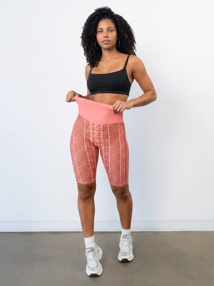 Woman pulling up the high-rise waistband of SunBorn N'Donso™ Biker Shorts in Adaeze Pink in a white studio