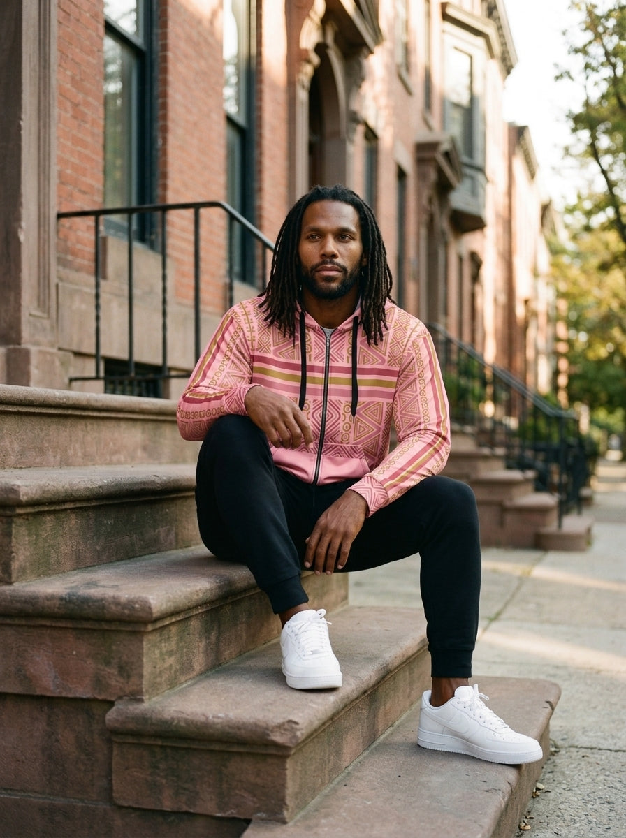 Man sitting on brownstone steps wearing the SunBorn N'Donso™ Zip Hoodie in Eze Pink with black joggers and white sneakers