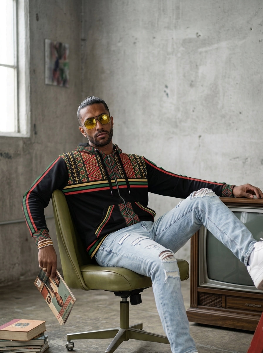 Man in yellow sunglasses sitting in a loft wearing the SunBorn N'Donso™ Zip Hoodie in Uhie Red with light ripped jeans