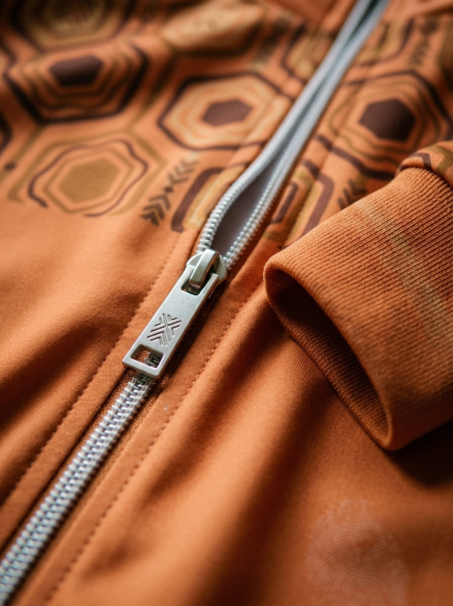 AnanseNova™ Statement Track Jacket in Sango Orange — close-up of the zipper pull, hexagon print and triple-stripe rib cuff on AkomaDrive™ brushed fleece