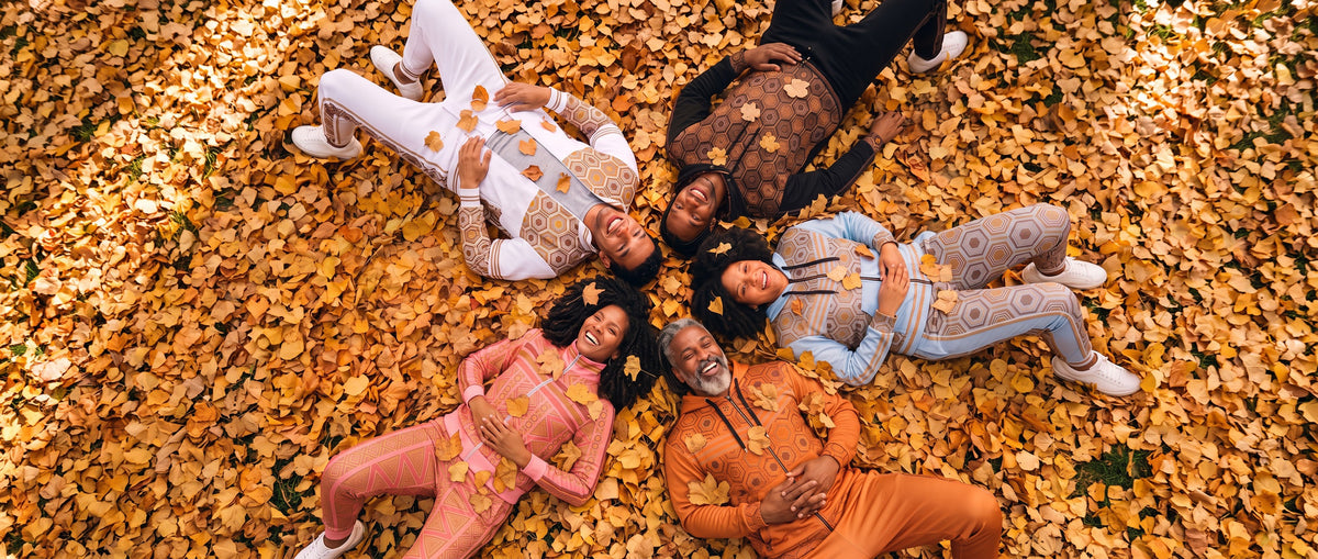 Five friends laughing as they lie in a pile of golden autumn leaves, wearing SunBorn tracksuits in white, black, light blue, orange and pink
