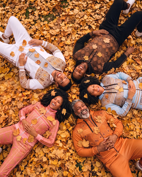 Five friends laughing as they lie in a pile of golden autumn leaves, wearing SunBorn tracksuits in white, black, light blue, orange and pink